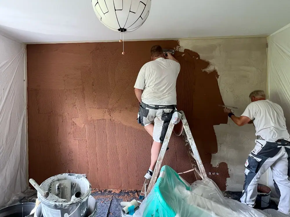 Zwei Maler tragen terrakottafarbene Spachtelmasse mit dem Zahnspachtel auf eine Wand auf